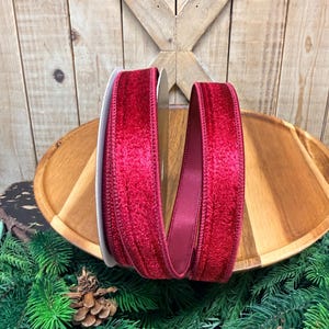 May include: A spool of deep red velvet ribbon with a textured, plush surface and stitched edges. The ribbon is wound around a central core, displaying a rich, saturated crimson color that contrasts with the smooth, satin-like finish on the inner side.
