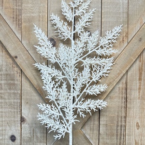 May include: A white, frosted artificial fern stem. The faux plant has a central stem with multiple branches of delicate, feathery leaves. The stem is against a rustic wooden background, suggesting a natural or farmhouse aesthetic.