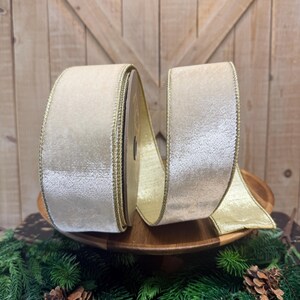 May include: A spool of wide, champagne-colored velvet ribbon featuring a metallic gold stitched edge. The ribbon has a soft, shimmering texture and is wound around a central core, with a portion of the fabric extending outward to display the finish.