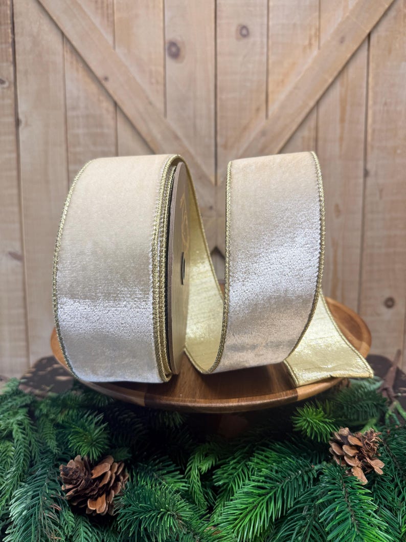 May include: A spool of wide, champagne-colored velvet ribbon featuring a metallic gold stitched edge. The ribbon has a soft, shimmering texture and is wound around a central core, with a portion of the fabric extending outward to display the finish.