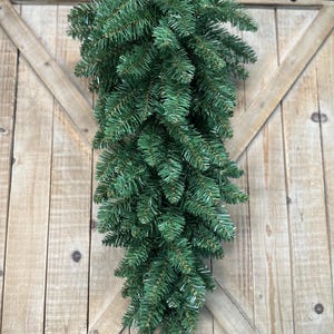 May include: A teardrop-shaped artificial evergreen swag featuring dense, realistic green pine needles. The foliage is arranged in a downward-tapering design, creating a full and lush appearance suitable for vertical display on a door or wall.