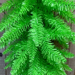 May include: Close-up of bright green artificial Christmas tree branches. The needles are a vibrant, solid green color, and the branches have a full, textured appearance. The background is a wooden fence.