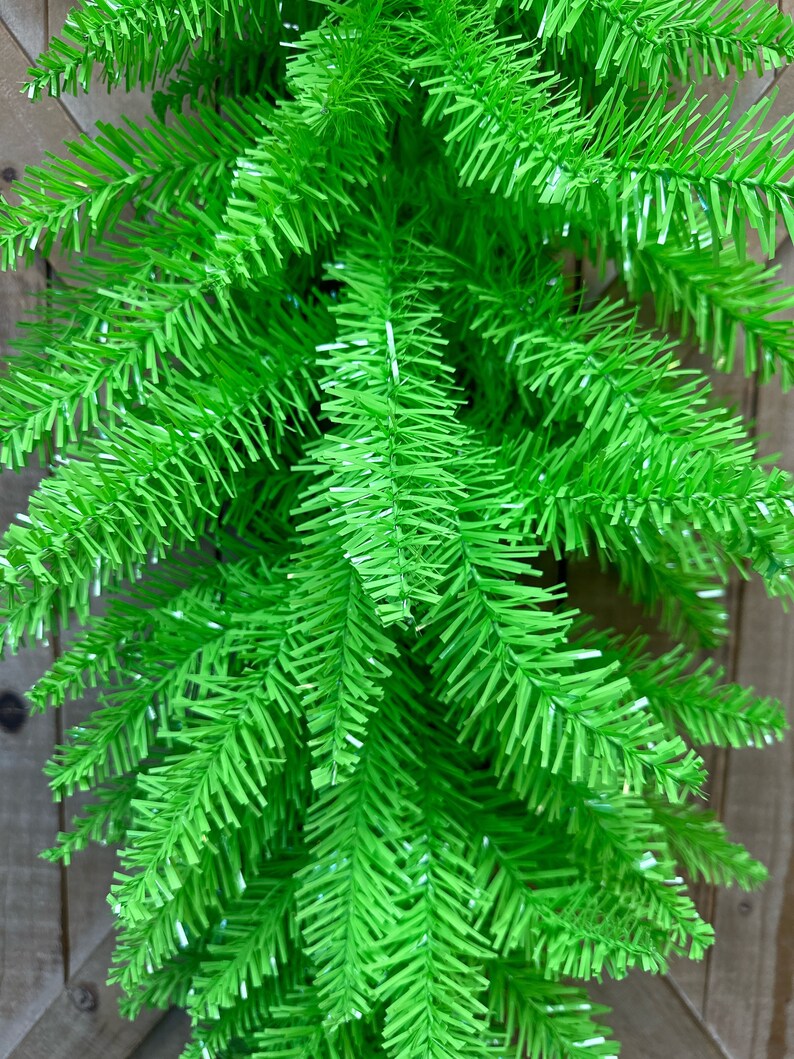 May include: Close-up of bright green artificial Christmas tree branches. The needles are a vibrant, solid green color, and the branches have a full, textured appearance. The background is a wooden fence.