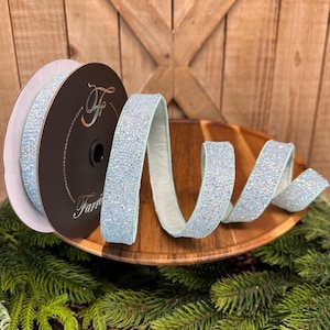 May include: A spool of light blue ribbon with a glittery, textured surface. The ribbon is displayed on a wooden tray with a green pine needle base. The ribbon is from the brand "Farri".