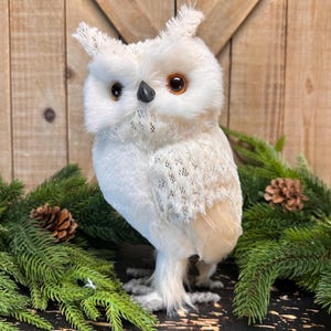 May include: A white plush owl figurine with brown eyes and a black beak. The owl has textured wings and is surrounded by green pine branches and pinecones. The background is a wooden surface.