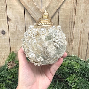 Pu&ograve; includere: Un addobbo natalizio sferico con base glitterata dorata, decorato con applicazioni in pizzo floreale color crema, grappoli di perle bianche artificiali e piccoli strass trasparenti. La parte superiore &egrave; rifinita con un tappo dorato metallizzato.