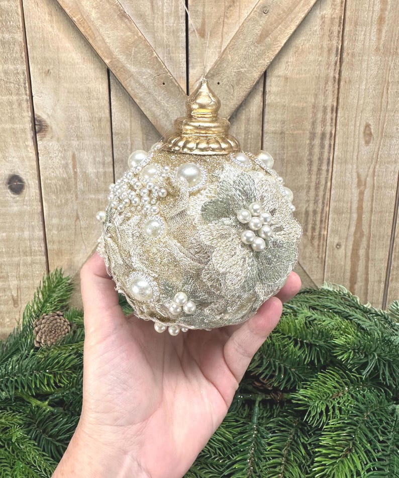 Pu&ograve; includere: Un addobbo natalizio sferico con base glitterata dorata, decorato con applicazioni in pizzo floreale color crema, grappoli di perle bianche artificiali e piccoli strass trasparenti. La parte superiore &egrave; rifinita con un tappo dorato metallizzato.