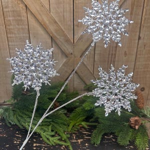 May include: Three silver snowflake ornaments on stems, perfect for holiday decor. Each snowflake is intricately detailed with sparkling crystals. The stems are silver and glittery, and the snowflakes are arranged against a backdrop of evergreen branches and a rustic wooden surface.