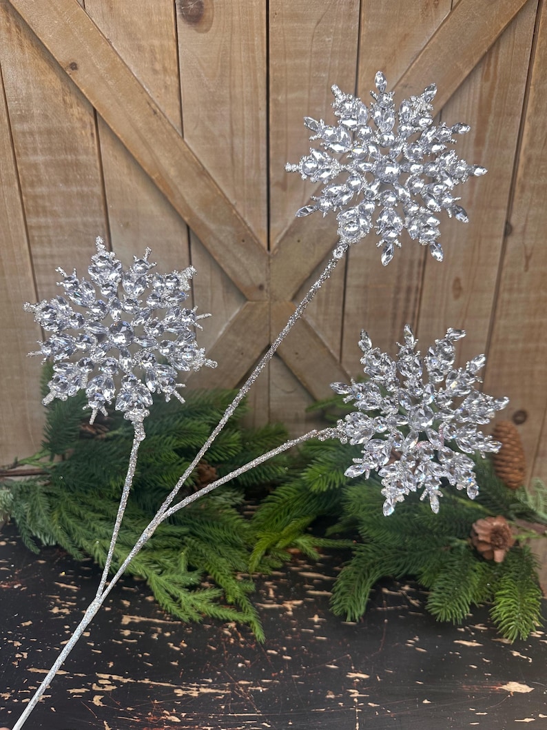 May include: Three silver snowflake ornaments on stems, perfect for holiday decor. Each snowflake is intricately detailed with sparkling crystals. The stems are silver and glittery, and the snowflakes are arranged against a backdrop of evergreen branches and a rustic wooden surface.