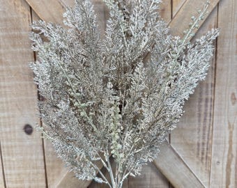 May include: A silver glittered artificial plant with multiple branches. The plant has a central stem and is surrounded by green pine needles and pine cones. The background is a wooden door.