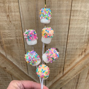May include: A skewer of marshmallow treats, each coated in colorful sprinkles. The marshmallows are white with a rounded top covered in a variety of pastel-colored sprinkles. The skewer is held in a hand against a wooden background.