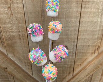 May include: A skewer of marshmallow treats, each coated in colorful sprinkles. The marshmallows are white with a rounded top covered in a variety of pastel-colored sprinkles. The skewer is held in a hand against a wooden background.