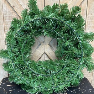 May include: A green artificial evergreen wreath with a wire frame. The wreath is on a black surface.