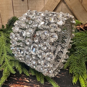 May include: A silver-toned, wire-framed decorative object encrusted with numerous clear, faceted crystals. The crystals vary in size and shape, creating a sparkling effect. The object is resting on a bed of green pine needles, suggesting a holiday theme.