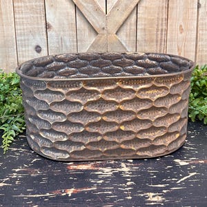 May include: An oval-shaped planter made of metal with a hammered finish. The planter is a dark brown color with a subtle gold sheen.