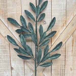 May include: A single artificial olive branch with multiple green leaves and a brown stem. The leaves have a textured, speckled appearance. The branch is set against a light brown wooden background.
