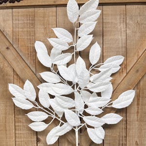 May include: A white artificial leaf stem with multiple textured leaves. The leaves are clustered along the stem, creating a full and decorative appearance. The stem is set against a wooden background.