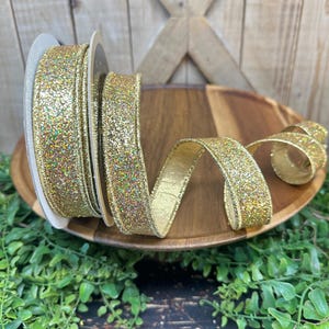 May include: A spool of gold glitter ribbon with a metallic sheen. The ribbon is displayed on a wooden tray, with a portion unrolled. The ribbon is approximately 1.5 inches wide. The background includes greenery and a wooden surface.