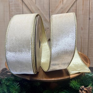 May include: A spool of wide, cream-colored velvet ribbon featuring a metallic gold edge. The ribbon is wound around a central core, with a portion of the fabric extending outward, showcasing the soft texture and the shimmering gold trim on both sides.