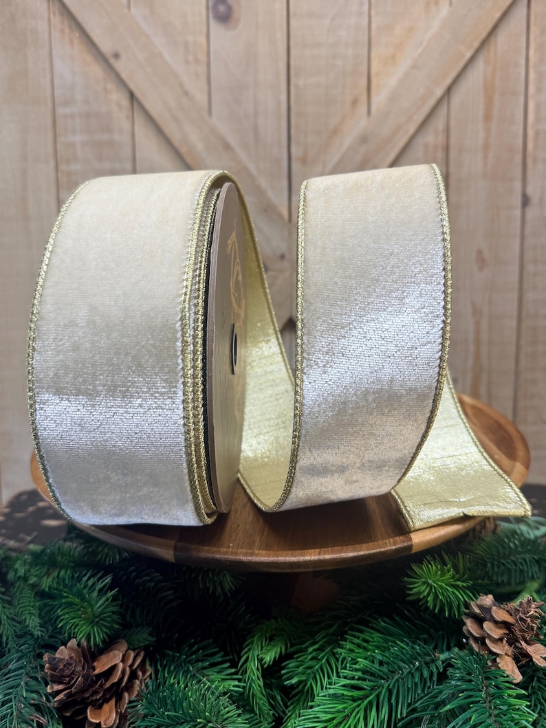May include: A spool of wide, cream-colored velvet ribbon featuring a metallic gold edge. The ribbon is wound around a central core, with a portion of the fabric extending outward, showcasing the soft texture and the shimmering gold trim on both sides.