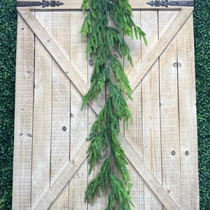 May include: A long, green garland of artificial pine branches hangs from a rustic wooden door with a diagonal X pattern. The door has two black metal hinges with decorative accents.