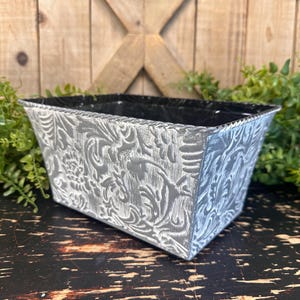 May include: A rectangular planter with a silver finish and embossed floral design. The planter has a black interior and is set against a wooden background. The planter is ideal for displaying plants.