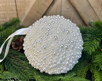 May include: A round, white Christmas ornament covered in small, shiny pearls. A white ribbon is attached for hanging. The ornament rests on a bed of green pine needles and a small pine cone, with a wooden background.