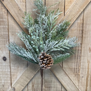 May include: A faux evergreen stem with a brown pine cone and green foliage. The artificial plant has a dusting of white snow and is set against a wooden background. The stem is ideal for seasonal decor.