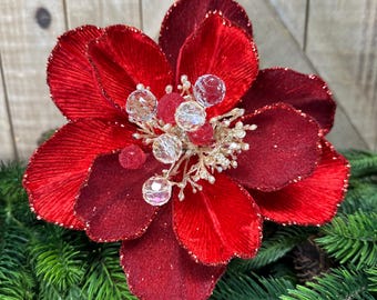 Può includere: Un fiore artificiale rosso scuro con petali testurizzati dai bordi glitterati. Il centro presenta rami dorati decorati con perline sferiche trasparenti sfaccettate e piccole bacche rosse, creando un elemento decorativo floreale.