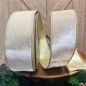 May include: A spool of wide, cream-colored velvet ribbon featuring a metallic gold edge trim. The ribbon has a soft, textured surface and is wound around a central core, with a portion of the shimmering gold-toned underside visible.