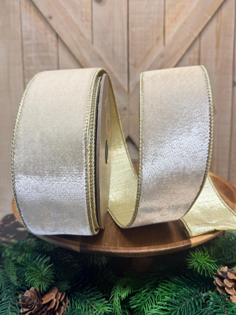 May include: A spool of wide, cream-colored velvet ribbon featuring a metallic gold edge trim. The ribbon has a soft, textured surface and is wound around a central core, with a portion of the shimmering gold-toned underside visible.