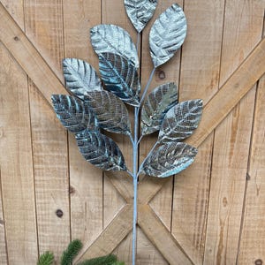 May include: A decorative metal leaf stem with silver and teal leaves. The leaves have a textured pattern and are attached to a light blue stem. The stem is displayed against a wooden background with greenery at the base.