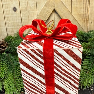 May include: A decorative Christmas gift box with a red velvet ribbon and bow. The box has a white and red striped pattern with glitter accents. A small gold bell ornament hangs from the bow. Green pine branches and a wooden background complete the festive scene.