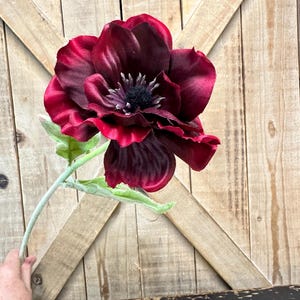 May include: A large, artificial burgundy flower with a dark center and green leaves. The flower has multiple layers of petals and is held by a light green stem. The background is a wooden surface.