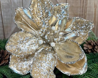 May include: A decorative artificial flower featuring metallic gold petals with silver glitter edges and accents. The center of the flower is embellished with small white faux pearls and gold glitter, creating a textured, multi-layered appearance.