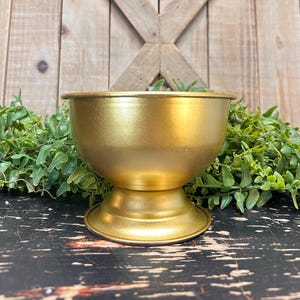 May include: A gold metal planter with a round bowl and a pedestal base. The planter is made of metal and has a shiny, metallic finish.