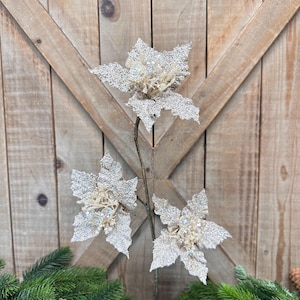 May include: Three glittery poinsettia flower stems with silver petals and a beige center. The flowers are arranged on a brown stem, with green pine branches and pine cones in the foreground. The background is a wooden door.