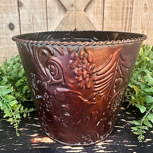 May include: A copper-colored metal planter with a decorative embossed floral design. The planter has a rope-like trim around the top edge.