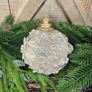 Pu&ograve; includere: Una decorazione natalizia sferica con un tappo metallico dorato e una superficie ricoperta da un intricato tessuto in pizzo dorato, arricchito da perle artificiali bianche di varie dimensioni e piccoli dettagli floreali argentati, appesa a un filo.