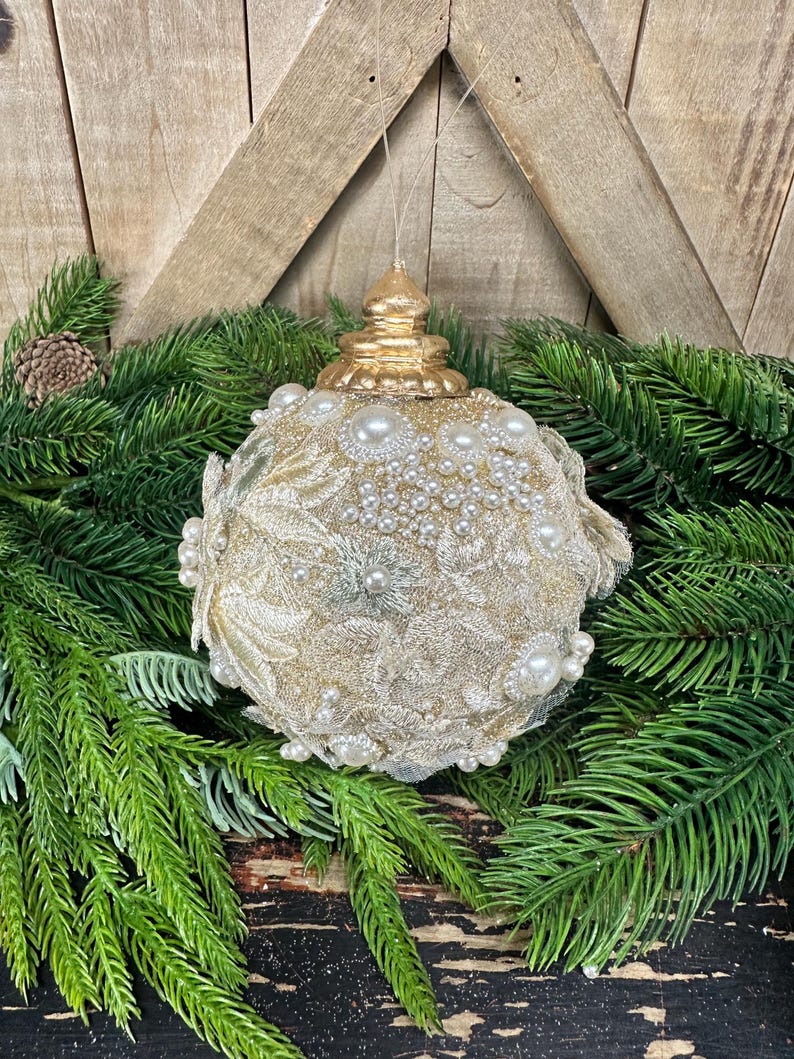 Pu&ograve; includere: Una decorazione natalizia sferica con un tappo metallico dorato e una superficie ricoperta da un intricato tessuto in pizzo dorato, arricchito da perle artificiali bianche di varie dimensioni e piccoli dettagli floreali argentati, appesa a un filo.