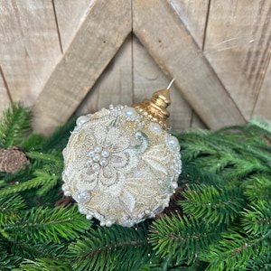 Pu&ograve; includere: Una pallina di Natale rotonda con intricati ricami floreali color crema e dettagli in filo dorato. La superficie &egrave; impreziosita da piccole perle artificiali bianche di varie dimensioni. Un tappo in metallo dorato si trova in cima con un occhiello.