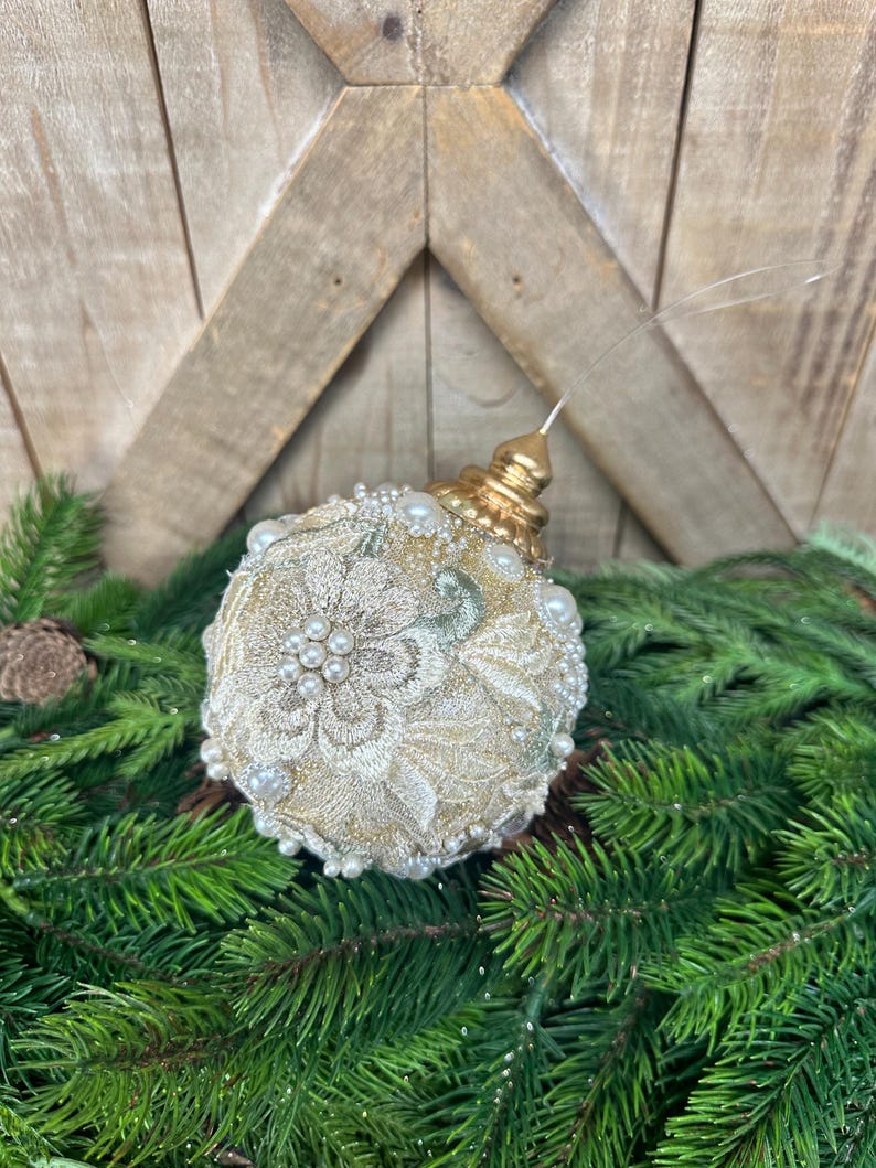 Pu&ograve; includere: Una pallina di Natale rotonda con intricati ricami floreali color crema e dettagli in filo dorato. La superficie &egrave; impreziosita da piccole perle artificiali bianche di varie dimensioni. Un tappo in metallo dorato si trova in cima con un occhiello.