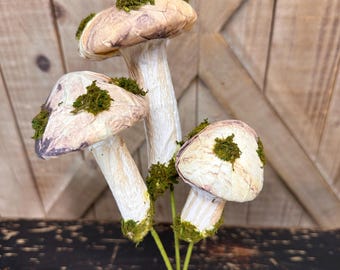 May include: A bouquet of three faux mushrooms with brown and white caps and green moss accents. The stems are green and the mushrooms are arranged in a bunch.