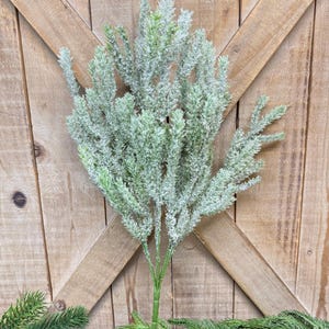 May include: A faux evergreen stem with a frosted appearance. The artificial plant has a green stem and branches, with a dusting of white, creating a wintery look. The stem is displayed against a wooden background, with additional evergreen branches at the base.