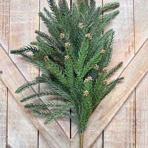 May include: A faux evergreen stem with green needles and small brown pinecones. The stem has a brown base and is set against a rustic wooden background. This artificial plant is suitable for seasonal decor.