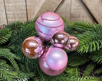 Puede incluir: Un grupo de adornos navideños rosas y oro rosa sobre un lecho de agujas de pino verdes. Algunos adornos tienen detalles de purpurina. El fondo es una superficie de madera.