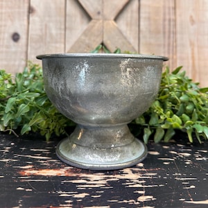 May include: A silver metal planter with a distressed finish. The planter has a round bowl shape with a pedestal base.