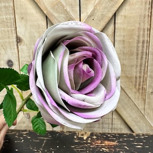 May include: A single artificial rose featuring layered petals in a gradient of muted grey, soft sage green, and vibrant purple. The flower is attached to a green stem with several small, pointed green leaves extending from the side of the stalk.