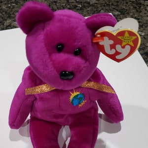 Puede incluir: Un oso de peluche morado Beanie Baby con una cinta dorada alrededor del cuello. El oso sostiene un pequeño globo terráqueo con el año 2000 bordado en él. El oso tiene una etiqueta Ty roja con las palabras "Beanie Original Baby" en ella.