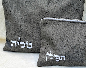 May include: Two gray fabric pouches with zippers, one larger than the other. The larger pouch has the word "שלום" embroidered on it in white. The smaller pouch has the word "דפלי" embroidered on it in white. Two black tefillin with gold accents are in the foreground.